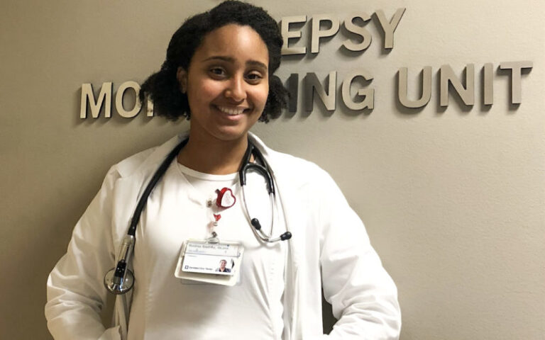 Andrea wearing medical uniform and stethoscope stands in front of hospital unit sign