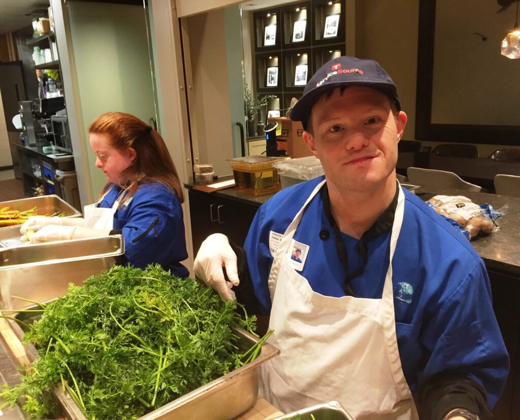 person in kitchen in front of salad greens smiling at camera